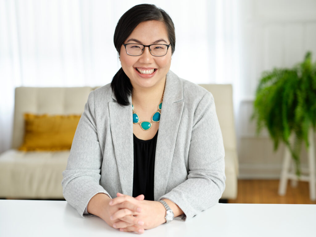 Picture of Councillor Tammy Hwang sitting at a white table with her hands crossed. She is wearing a grey blazer over a black shirt, with a teal necklace. She is wearing her hair tied back in a ponytail that is draping on the side of her right shoulder. She si smiling and inviting. 